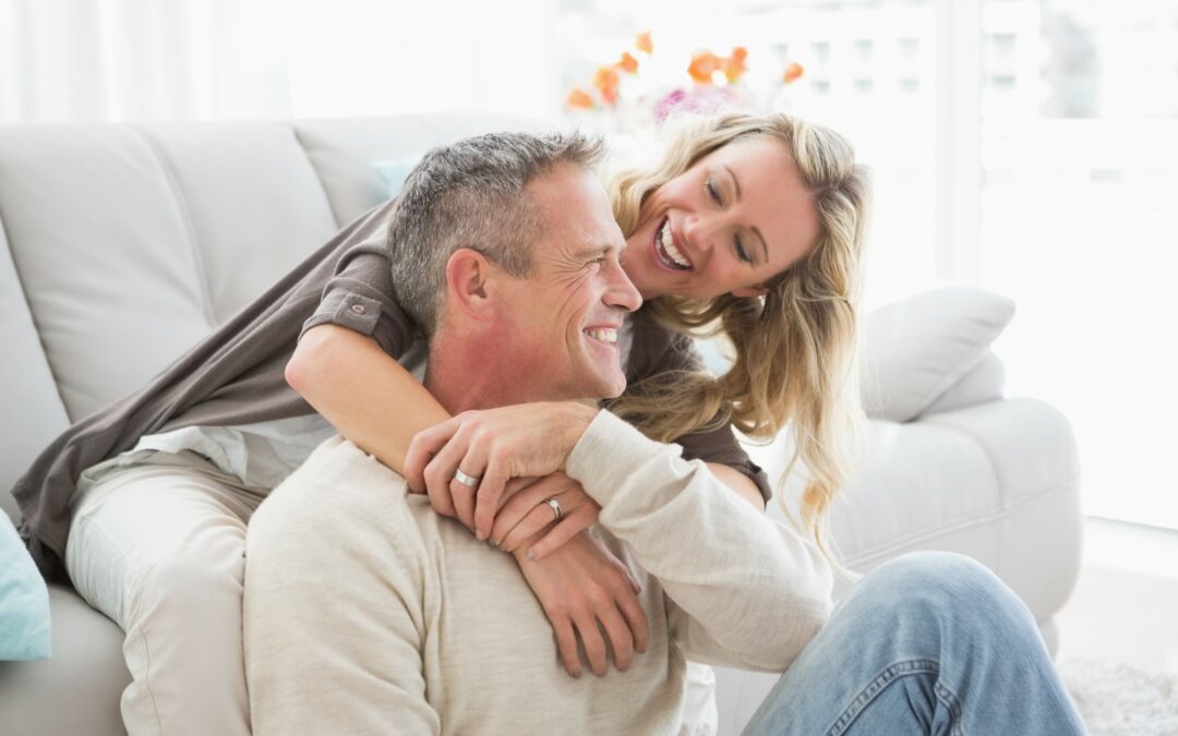älteres Pärchen kuschelt an einer Couch und blickt sich lächelnd an - die emotionale Intimität ist spürbar