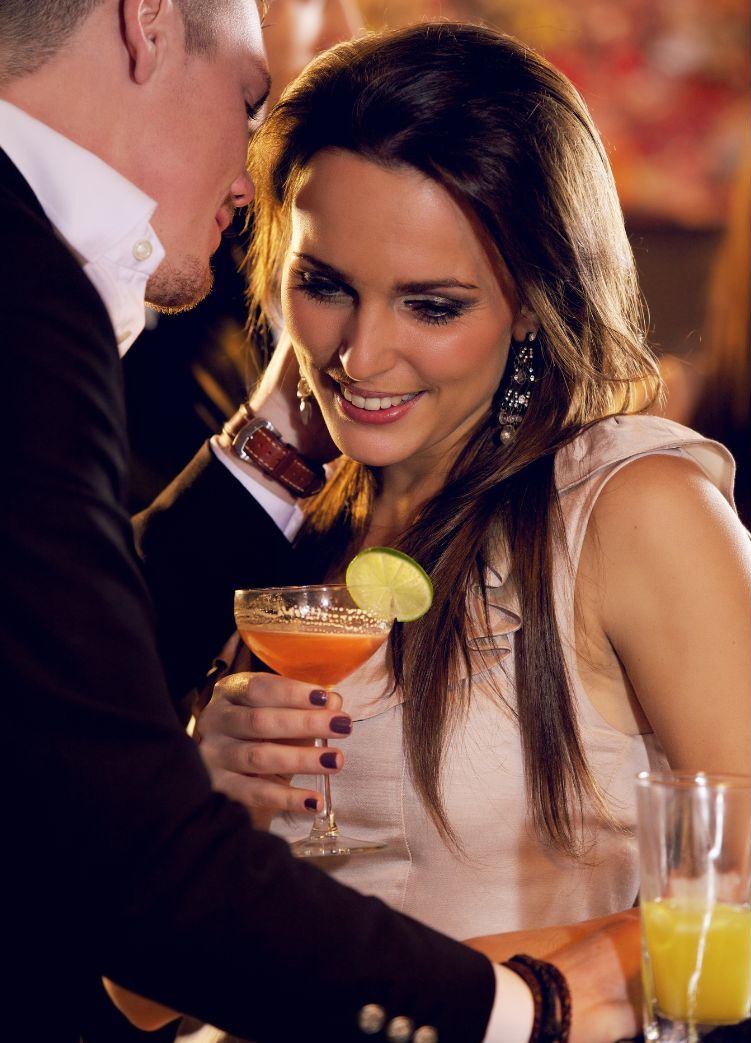 paartherapie online für glückliche partnerschaft Mann flüstert Frau ins Ohr, sie lächelt und hält ein Cocktailglas in der Hand - die Paartherapie online scheint zu funktionieren